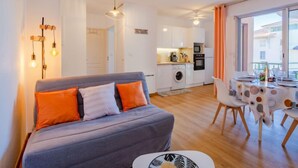 A modern apartment with a grey sofa, a dining area with a table and chairs, a kitchen with white cabinets, and a large window offering a view. - Quartier Latin by Interhome (Fréjus)