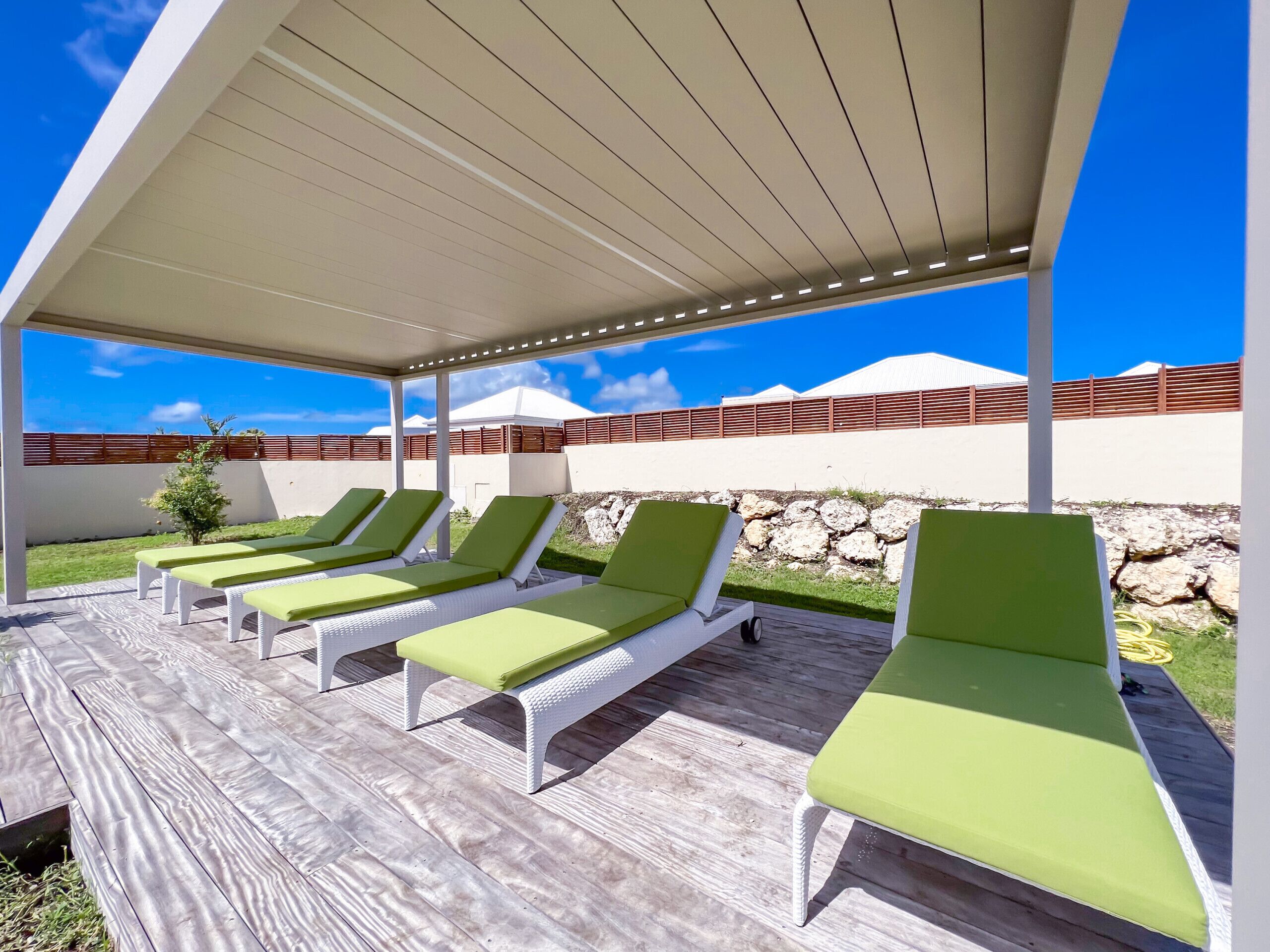 A covered outdoor seating area with white lounge chairs and green cushions, a wooden deck, and a view of a white building and a stone wall.