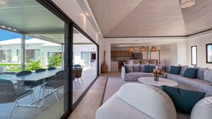Een moderne woonkamer met een grote bank, een salontafel en een eethoek met stoelen en een tafel. Achtergrond toont een keuken met een eettafel en stoelen.