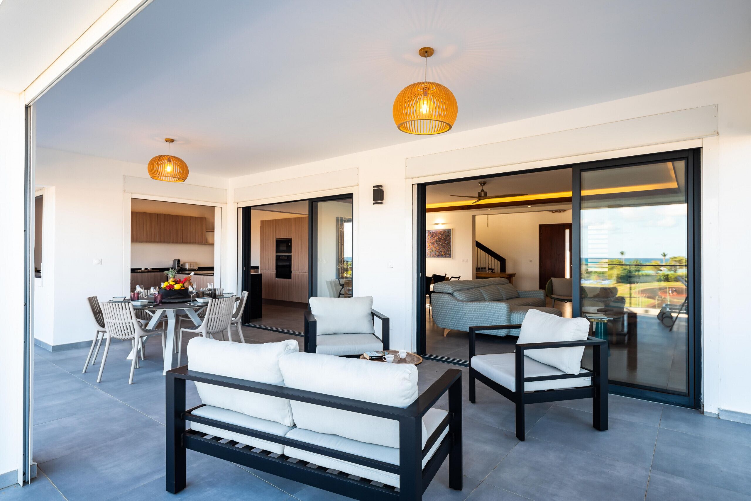 A modern living area with a sofa, chairs, and a dining table. There is a kitchen in the background and a sliding glass door leading to a balcony.