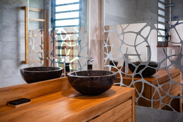 Un baño moderno con dos lavabos oscuros sobre un mueble de madera, un separador de vidrio y vistas a una zona de piscina.