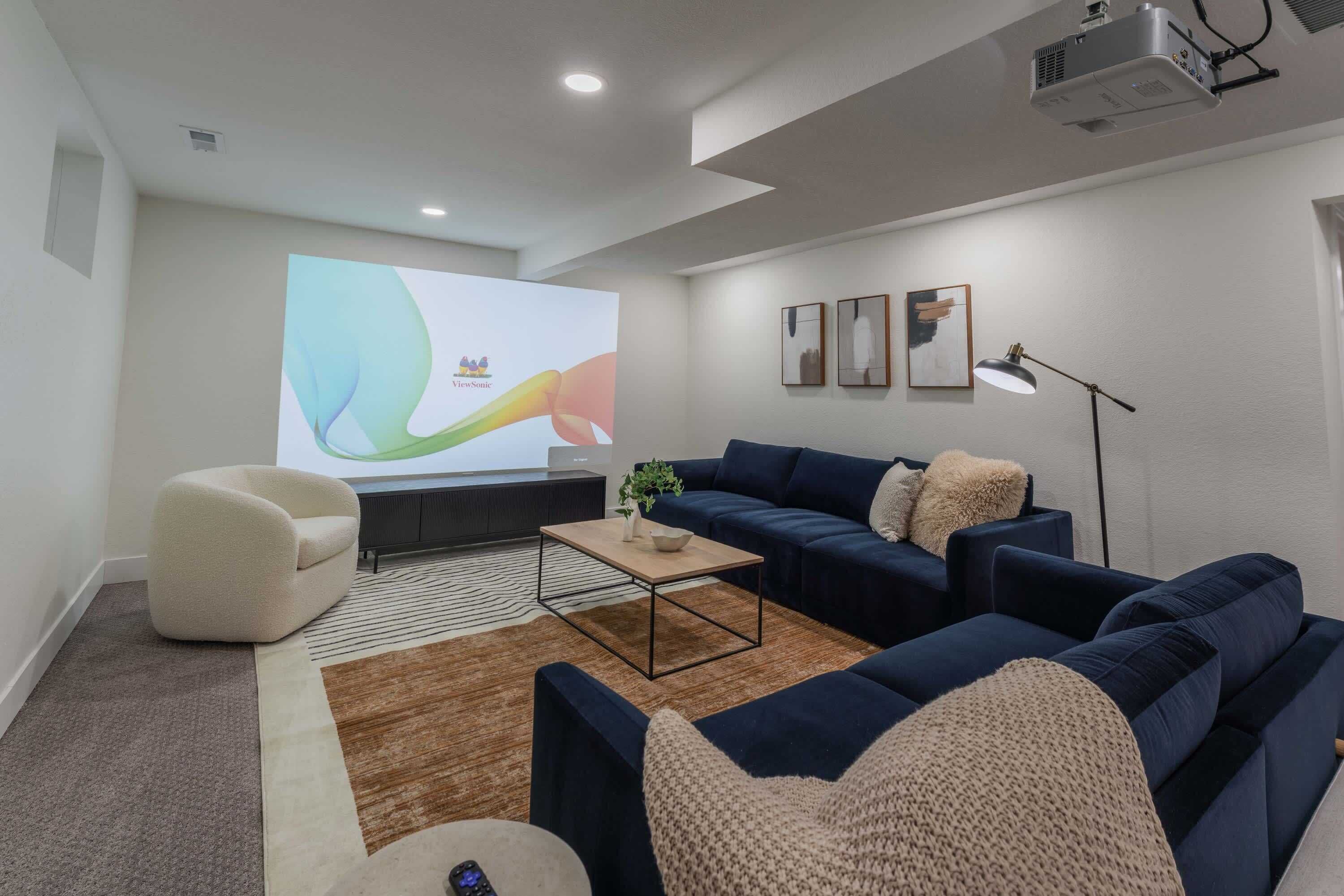 Ein modernes Wohnzimmer mit einem großen Bildschirm, dunkelblauen Sofas, einem weißen Sessel, einem Couchtisch und einer Stehlampe.