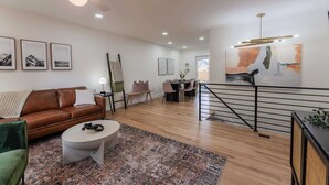 A modern living room with a brown leather sofa, a round coffee table, and a patterned rug. There is a dining area with a table and chairs, and a staircase with a railing. - Game Room- Hot Tub- Sauna & Projector! (Arvada)
