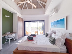 Une chambre à coucher comprenant un lit, un bureau, une chaise, un ventilateur de plafond, un tableau et une vue sur l’extérieur, accessible par une porte coulissante.