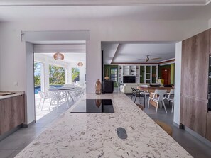 Une cuisine moderne avec un plan de travail en marbre, un coin repas avec des chaises blanches et une vue sur une piscine à l’extérieur.