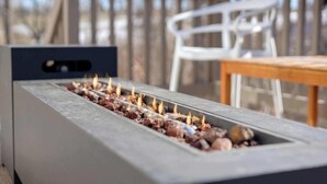 A modern outdoor fire feature with a metal frame and a rectangular stone base, surrounded by a wooden deck and white chairs. - Spacious home with Game Room, Hot Tub & Sauna! (Westminster)