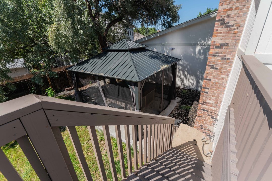 A covered patio area with a wooden railing, a brick wall, and a large tree providing shade.