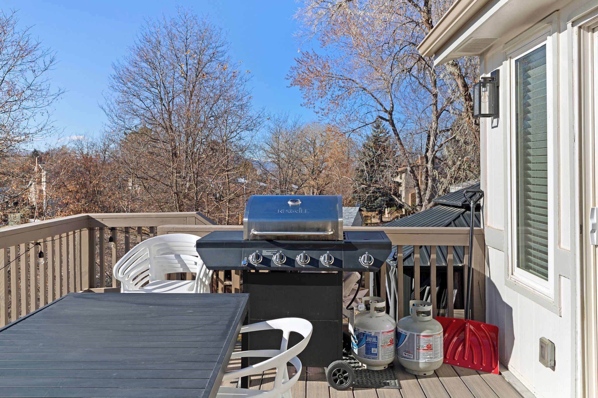 Un barbecue installé sur une terrasse, avec une table et des chaises, une bouteille de gaz et une pelle rouge.