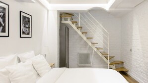 Une chambre avec un lit blanc, des murs blancs, un mur en briques blanches, un radiateur blanc et un escalier en bois.
