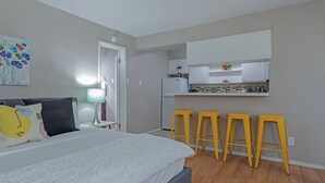 Quarto com cama, mesa de cabeceira, luminária, pintura e uma cozinha compacta com banquetas.