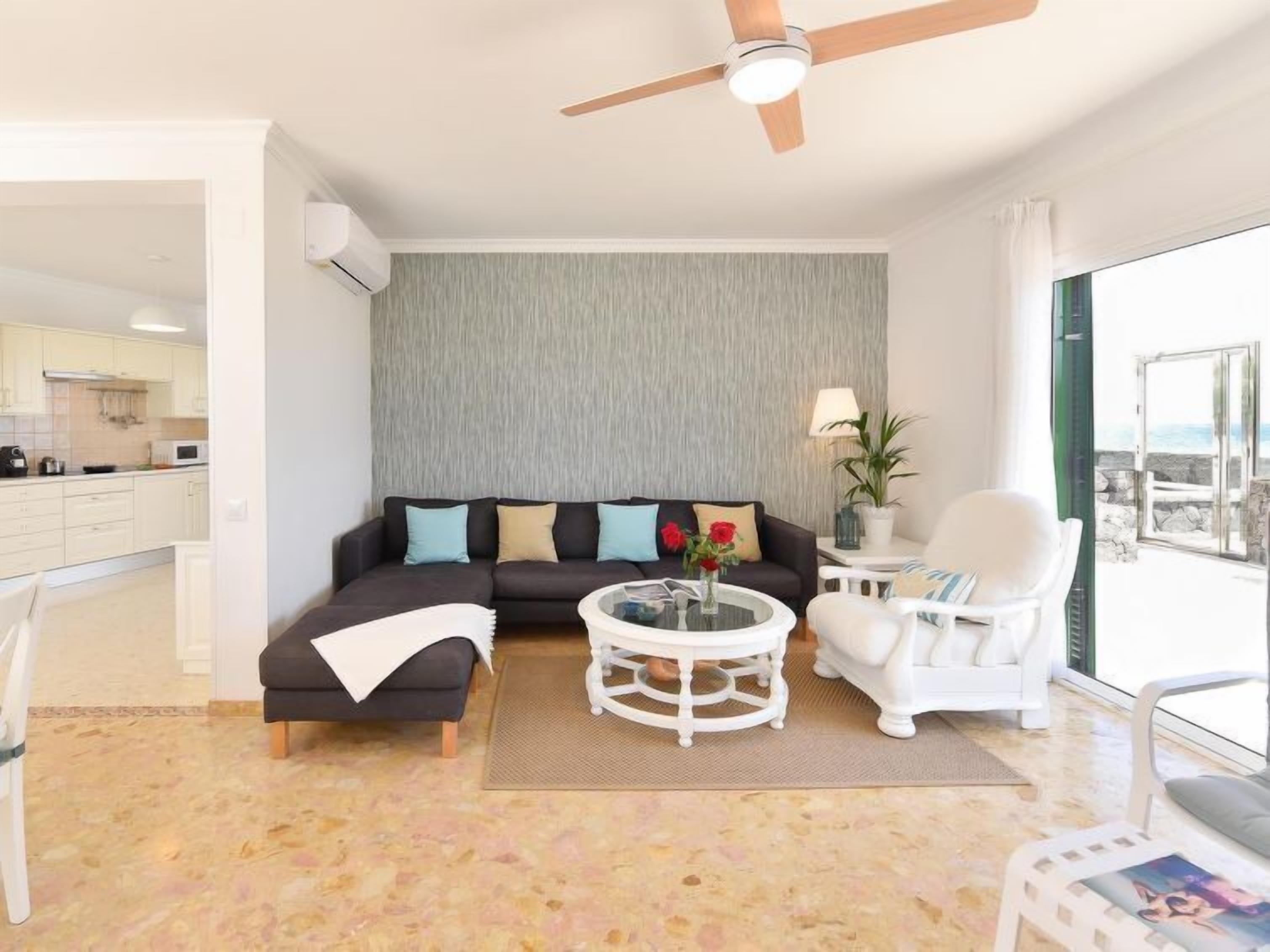 A modern living room with a sectional sofa, a round coffee table, and a white armchair. There is a view of the ocean through large windows.