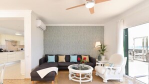Un salon moderne avec un canapé d’angle, une table basse ronde et un fauteuil blanc. De grandes fenêtres offrent une vue sur l’océan.