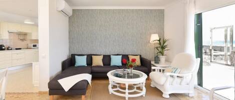 A modern living room with a sectional sofa, a round coffee table, and a white armchair. There is a view of the ocean through large windows.