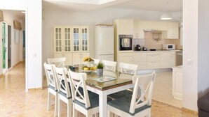 Une cuisine moderne avec des meubles blancs, un coin repas doté d’une table en verre et de chaises blanches, et un réfrigérateur visible.