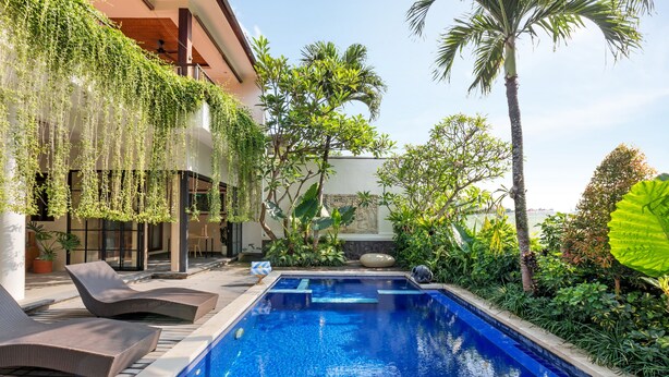 Une piscine entourée de chaises longues, de verdure luxuriante et d’un bâtiment doté d’un balcon.