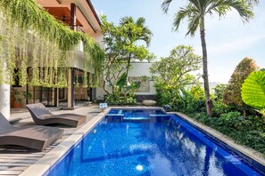 A swimming pool with lounge chairs, surrounded by lush greenery and a building with a balcony. - Kerta Mani Canggu (Canggu)
