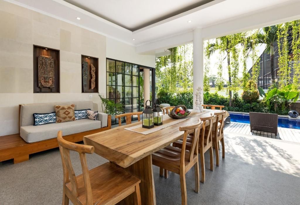 A modern dining area with a wooden table and chairs, a sofa with patterned cushions, and a view of a swimming pool and greenery.