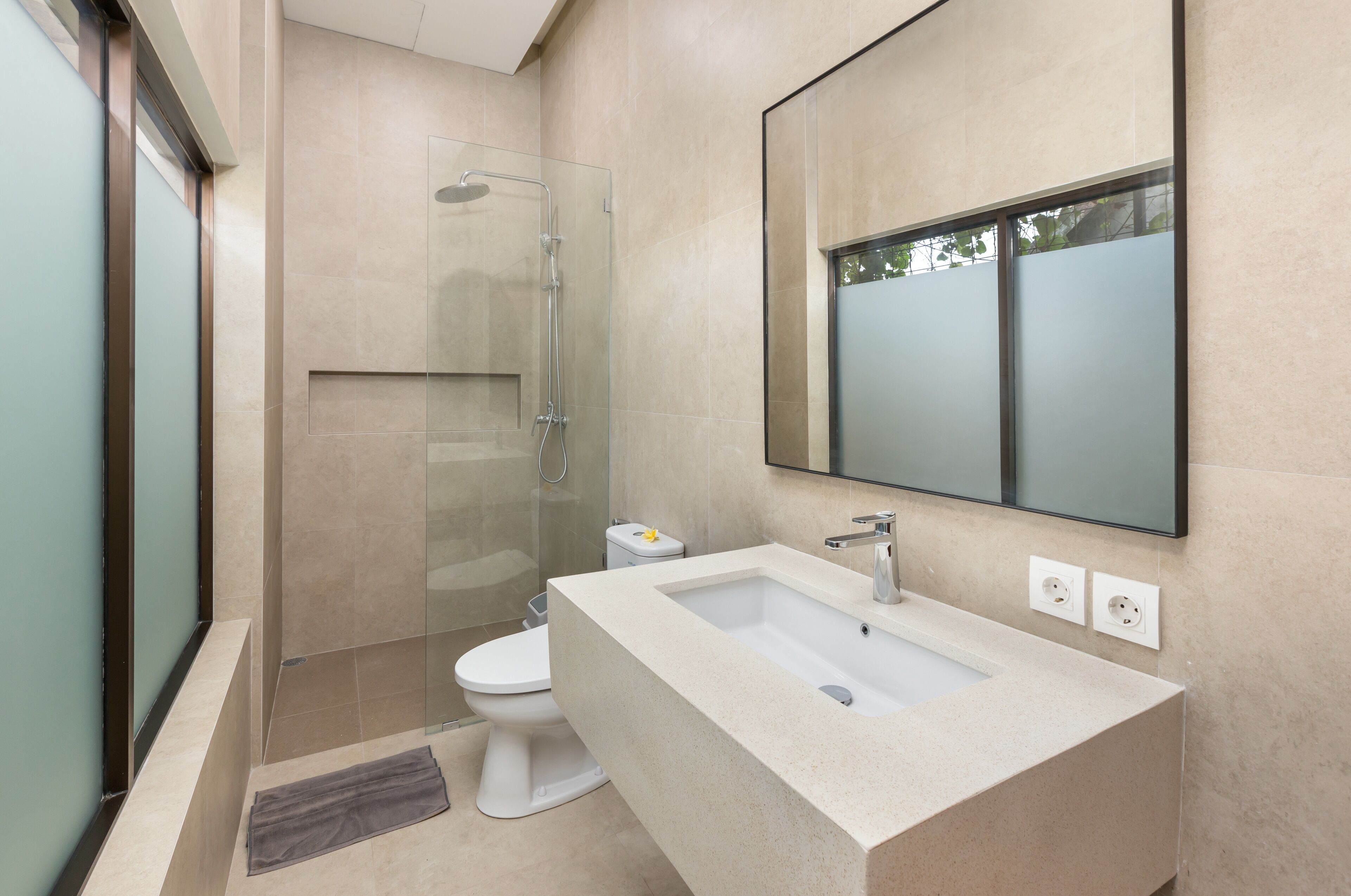 Un baño moderno con cabina de ducha de vidrio, inodoro blanco y lavamanos color beige con un espejo grande colgando sobre él.