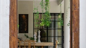 Un passage surmonté d’un cadre en bois mène à un coin repas comprenant une table et des chaises, des plantes suspendues et une grande fenêtre.