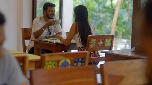 Una mesa de madera con una persona sosteniendo una taza, otra persona sentada al otro lado y una vista de follaje afuera.