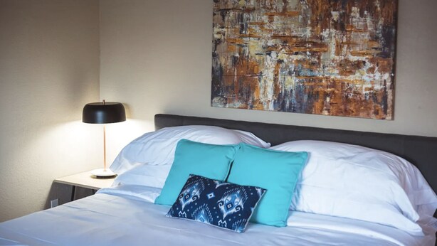 A neatly made bed with white linens and two teal pillows, a black lamp on a bedside table, and a large abstract painting on the wall. - Montrose Relaxing Studio! Medical Center 37 (Houston)