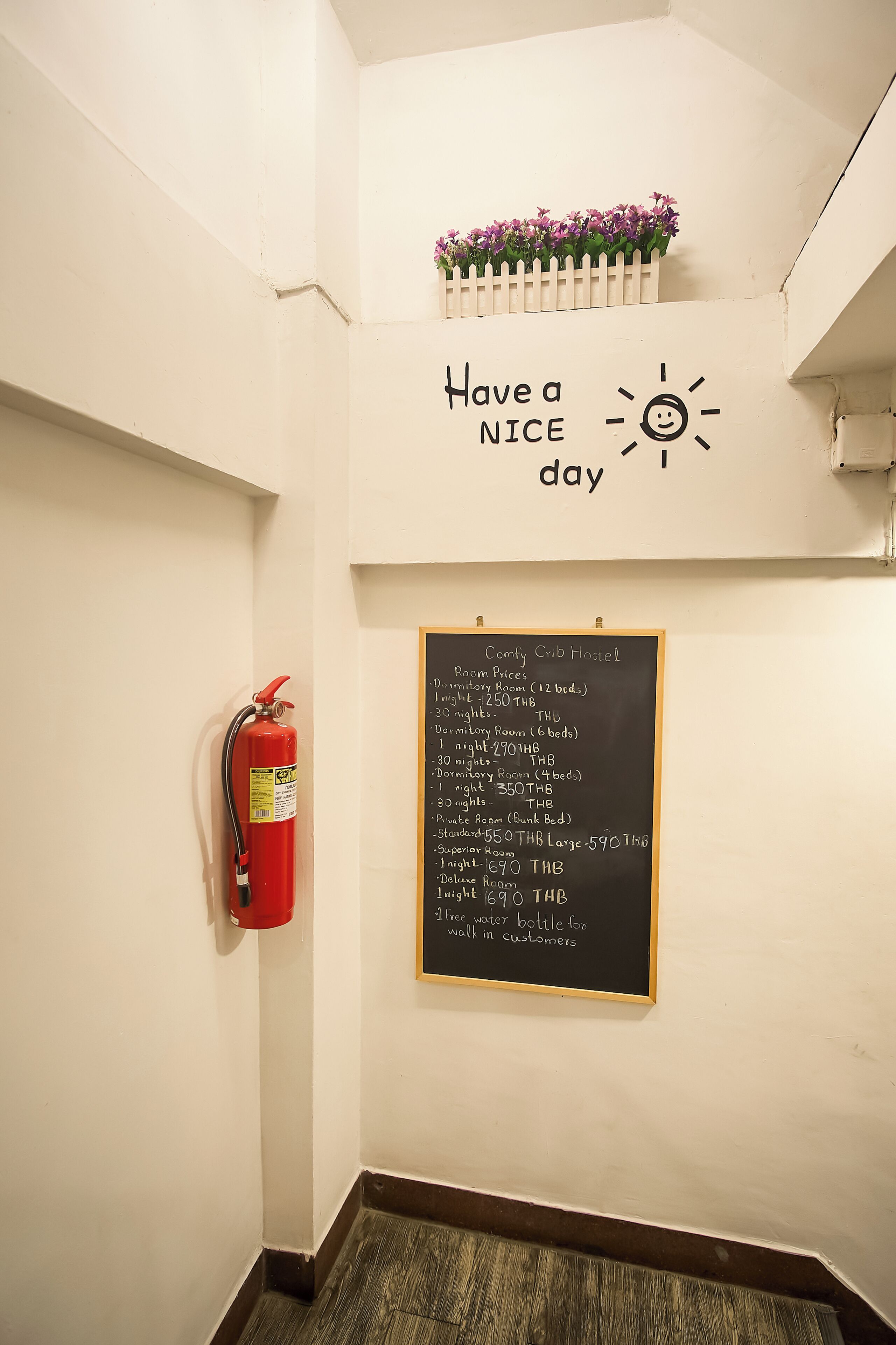A wall with a fire extinguisher, a chalkboard with room prices, and a sign saying 'Have a nice day' with a sun and smiley face.
