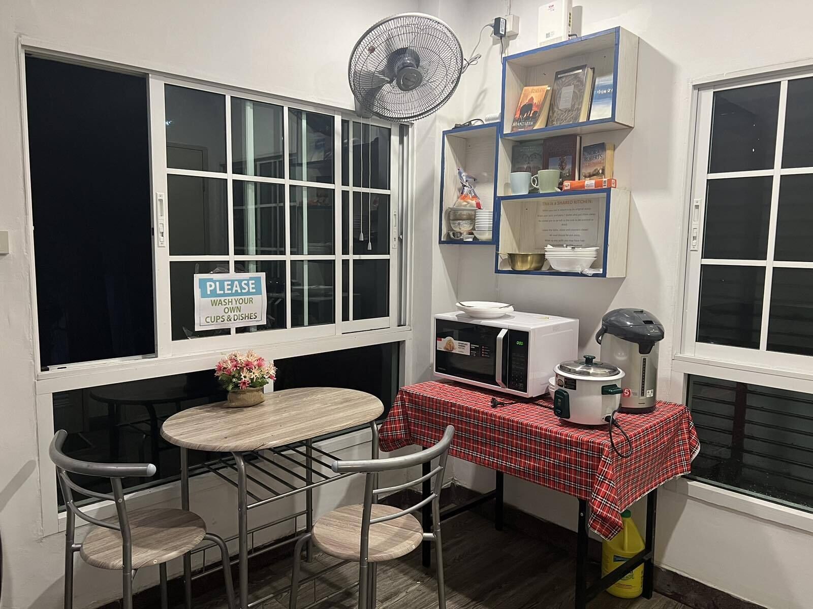 Una pequeña cocina americana equipada con microondas, hervidor de agua y ventilador. Hay un cartel solicitando a los huéspedes que laven sus propios vasos y platos.