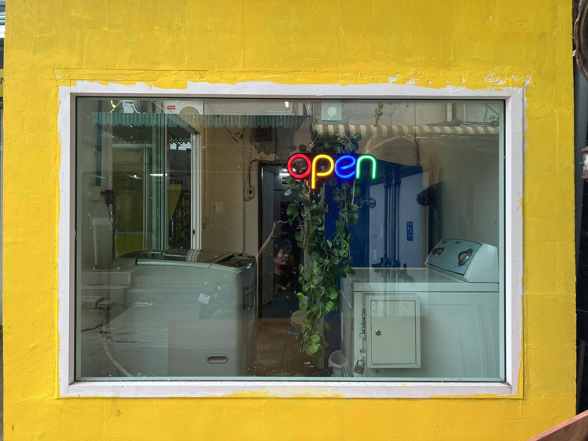 Une fenêtre avec une enseigne néon indiquant « ouvert », une porte vitrée et une petite plante à l’intérieur.