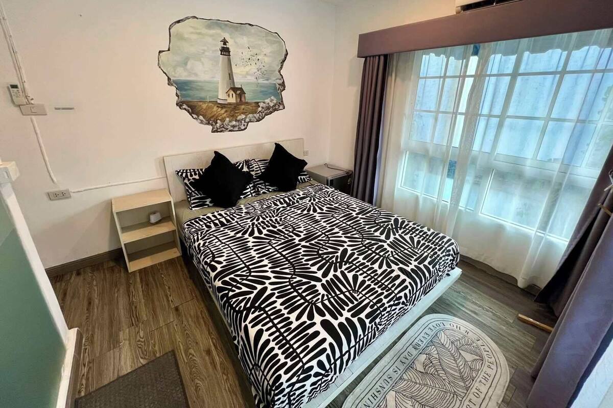 A bedroom with a bed, a nightstand, a wall-mounted artwork of a lighthouse, and a large window with curtains.