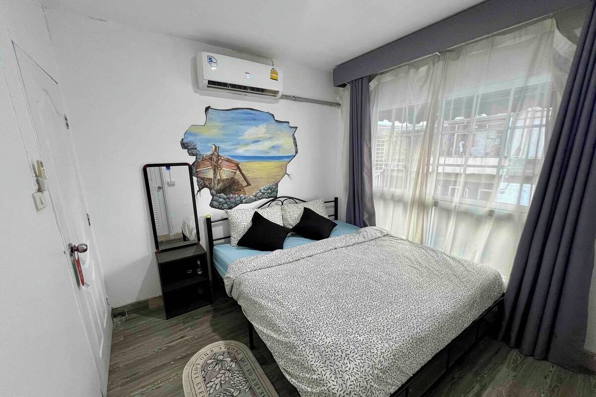 A bedroom with a bed, a nightstand, an air conditioner, a window with curtains, and a wall with a painted design.
