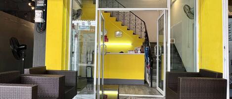A hostel interior with wicker furniture, a reception desk, and a staircase leading to upper floors.