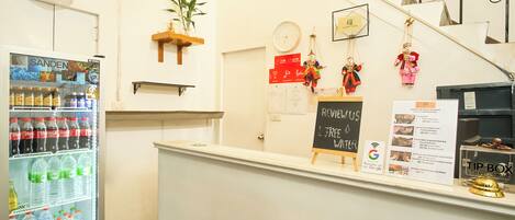 Zona de recepción de hotel con un refrigerador lleno de bebidas, un reloj y una pizarra con información sobre las habitaciones.
