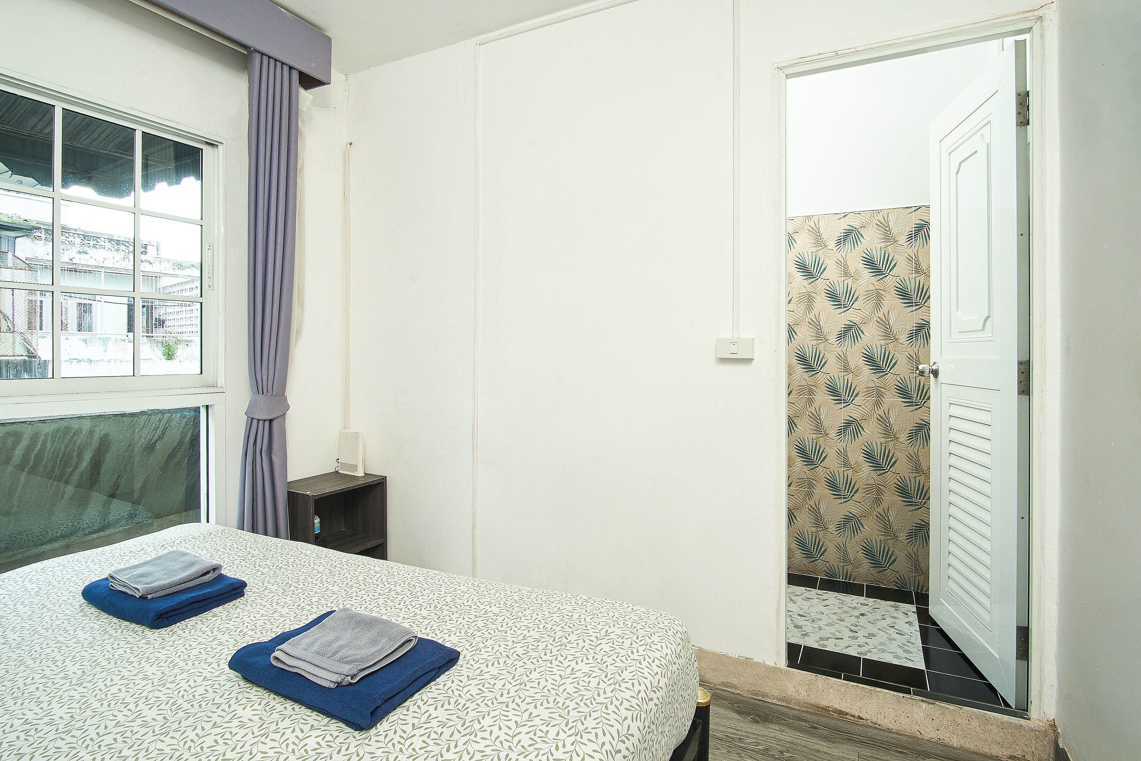 Ein Hotelzimmer mit Bett, Nachttisch, einem Fenster mit Vorhängen und einer Tür zu einem Badezimmer mit gemusterten Fliesen.