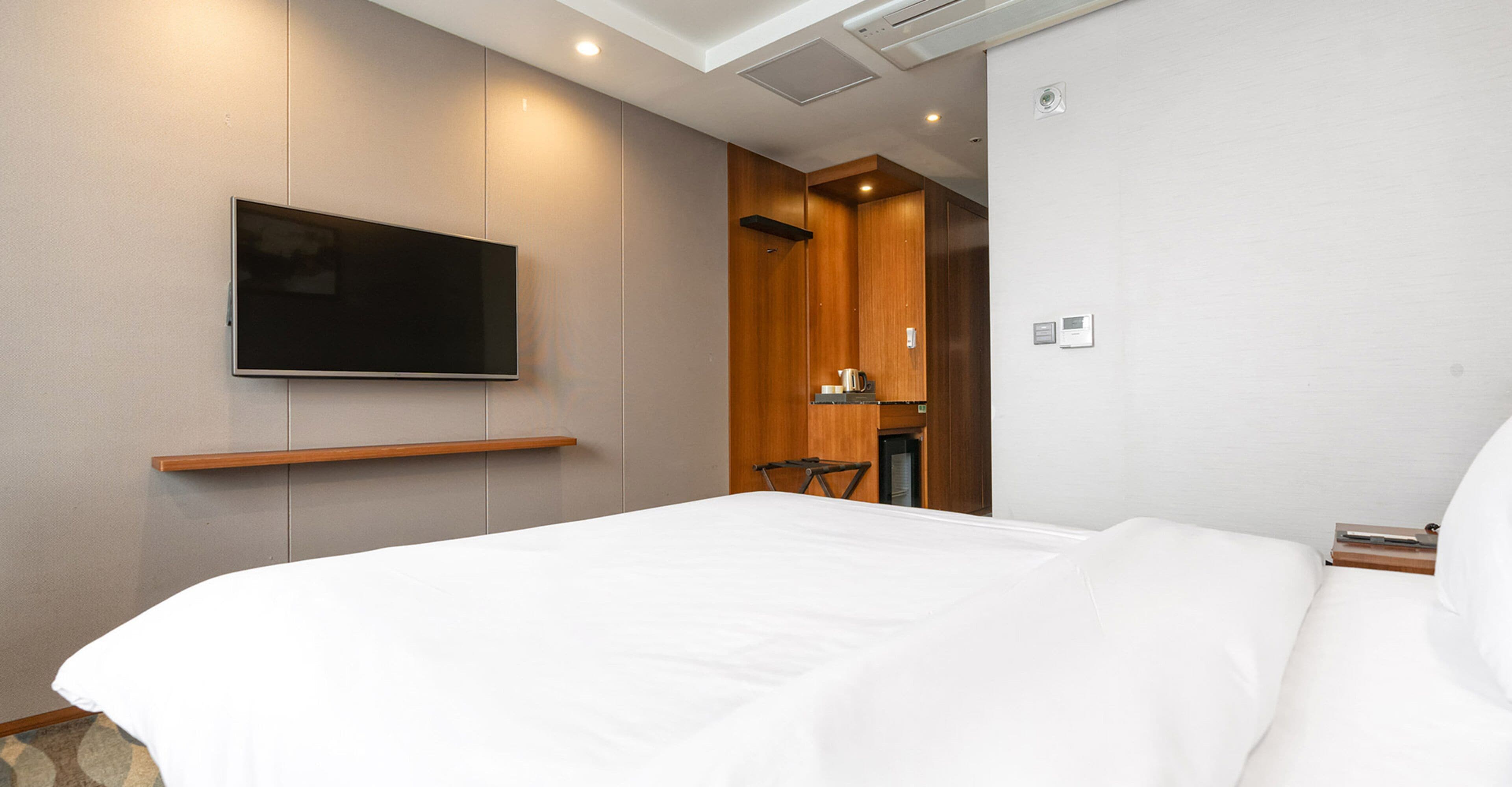 Habitación de hotel con una cama grande, un televisor montado, un armario de madera y una mesita con una tetera.