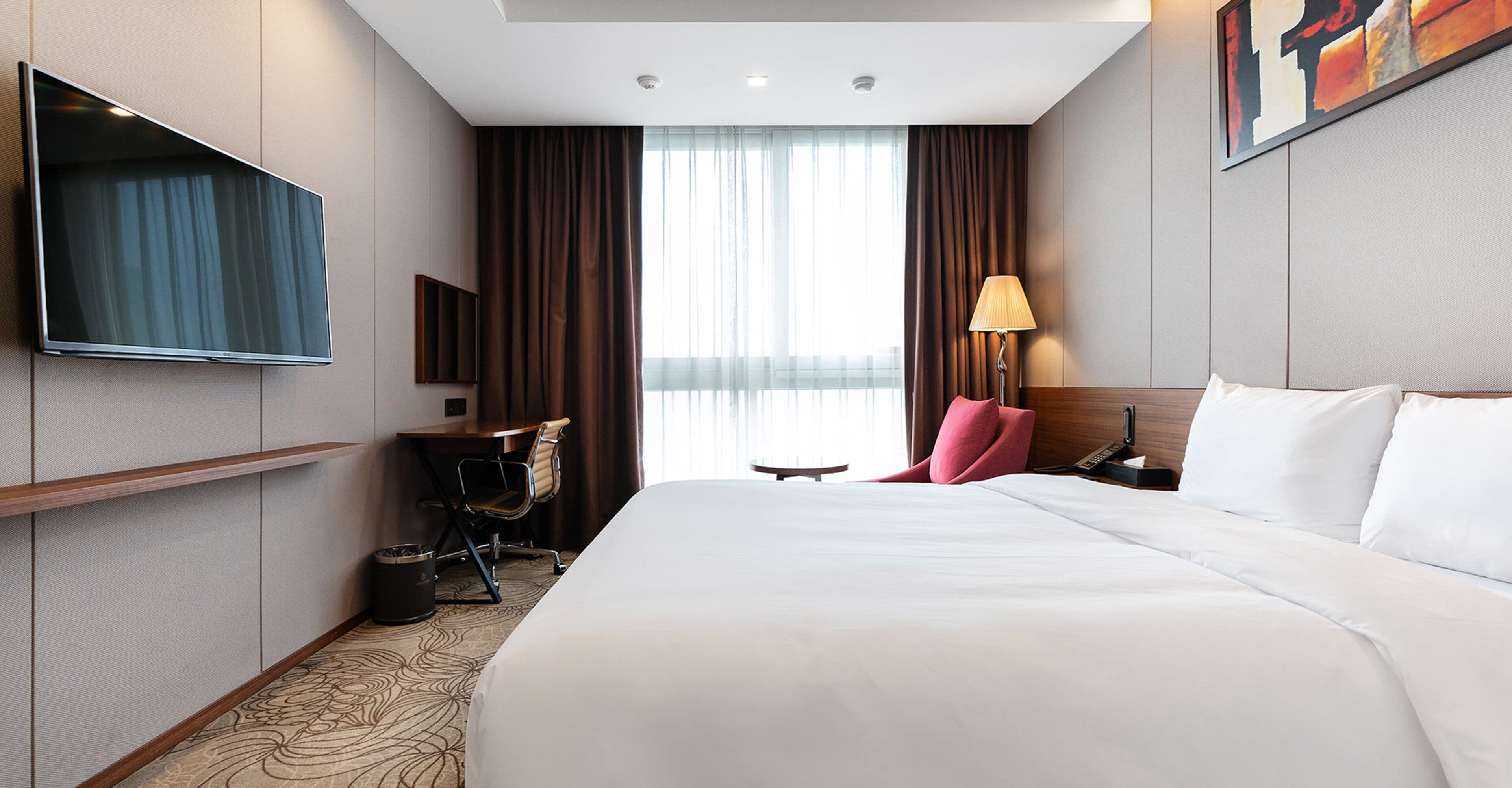 A hotel room with a large bed, a desk, a chair, a TV mounted on the wall, and a window with curtains.