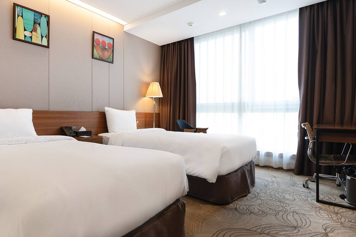 Une chambre d’hôtel avec deux lits, un bureau, une chaise, une lampe et des œuvres d’art au mur.