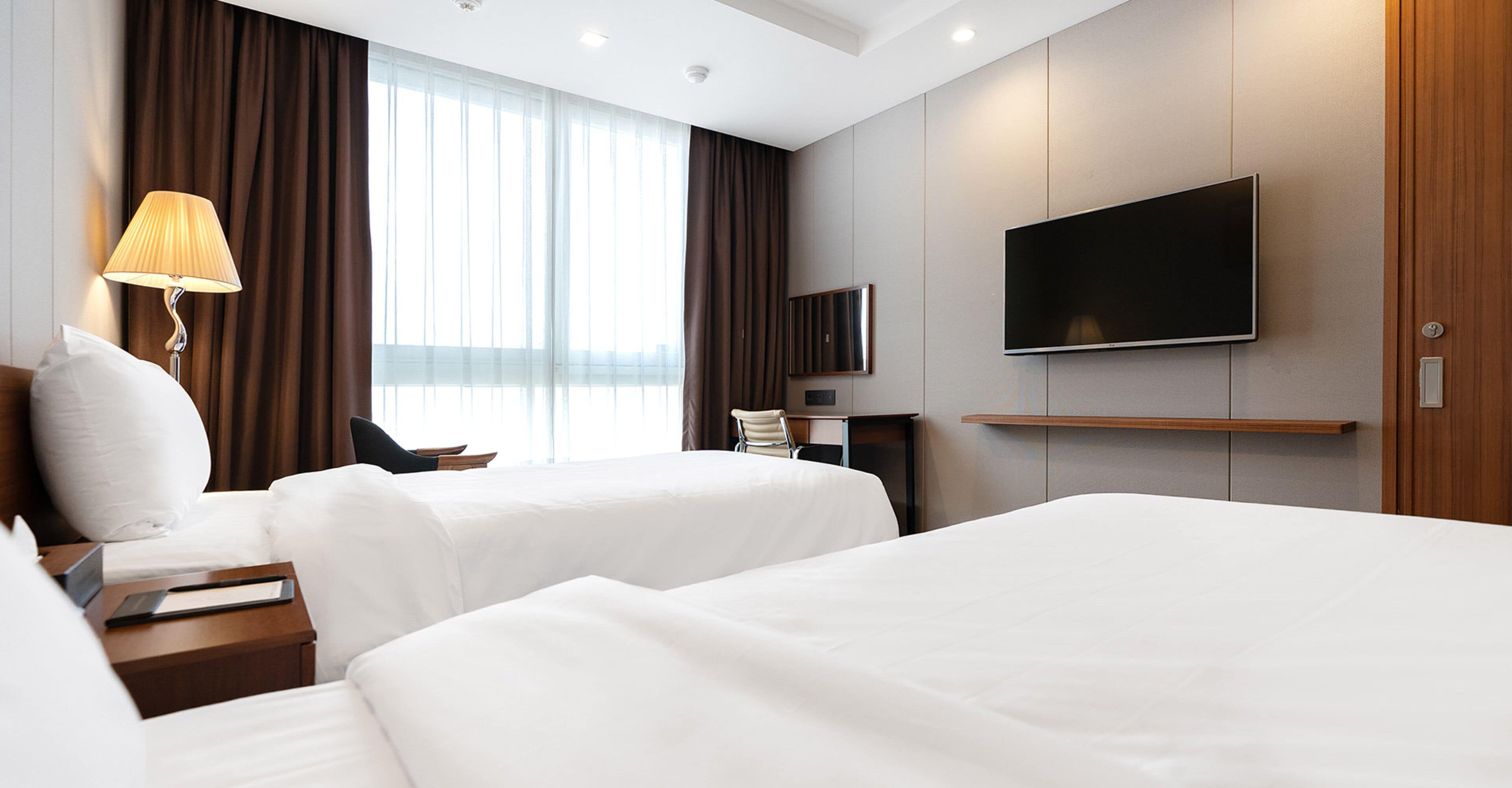 A hotel room with two beds, a TV on the wall, a lamp, and a window with curtains.