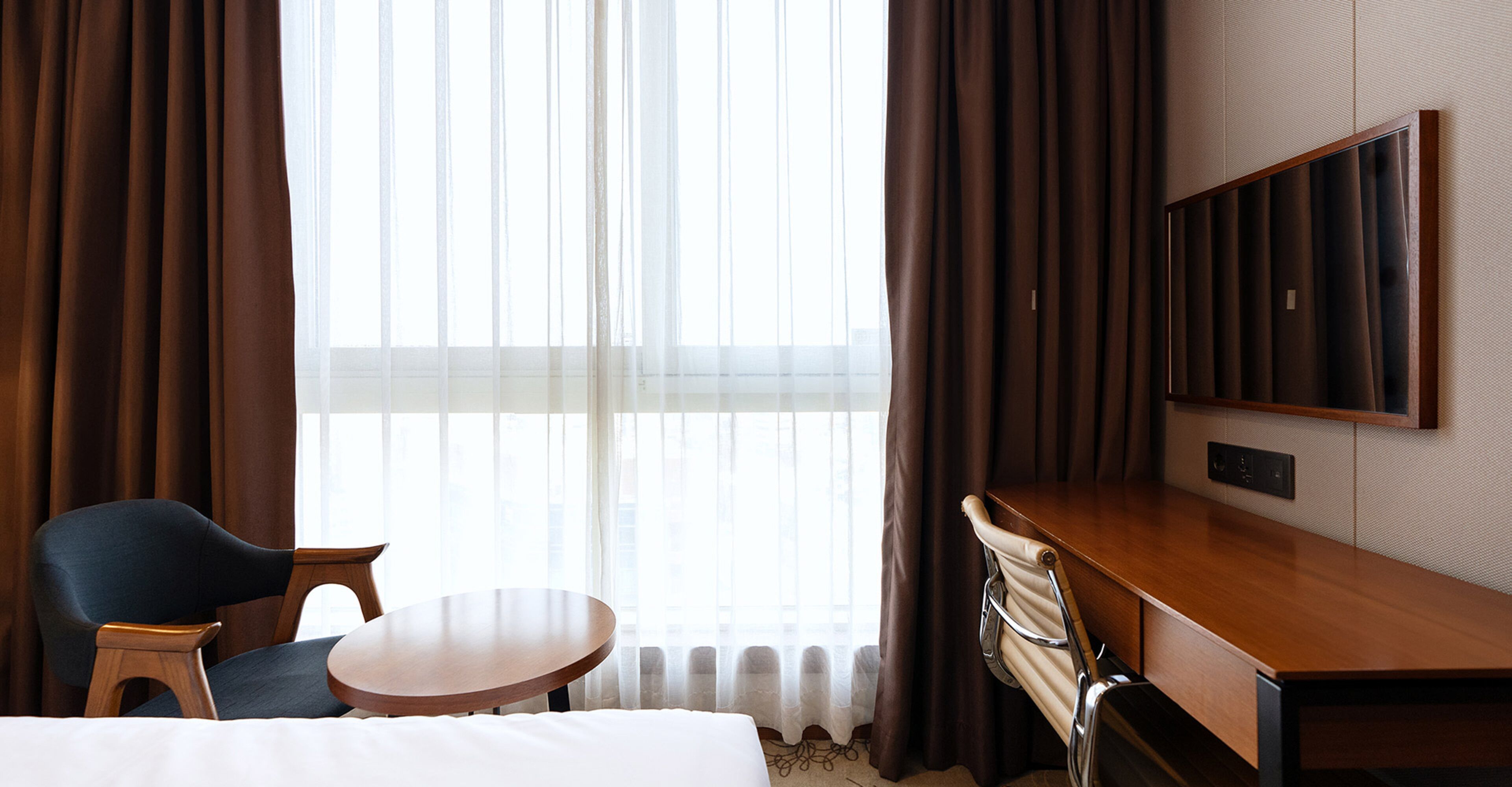 A hotel room with a bed, a desk, a chair, a round table, and a TV.