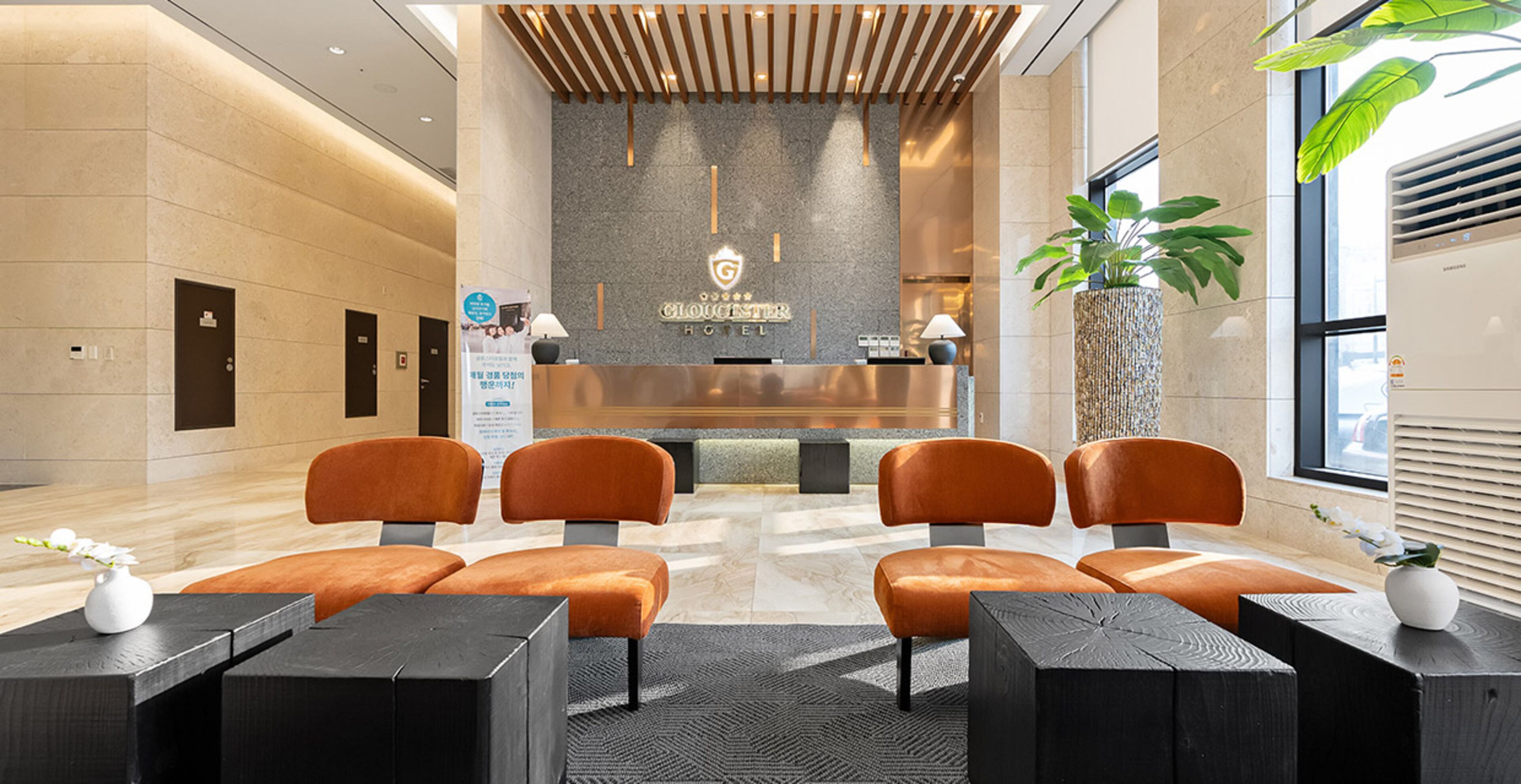 A hotel reception area with a modern design, featuring a reception desk, several orange chairs, and a large potted plant.