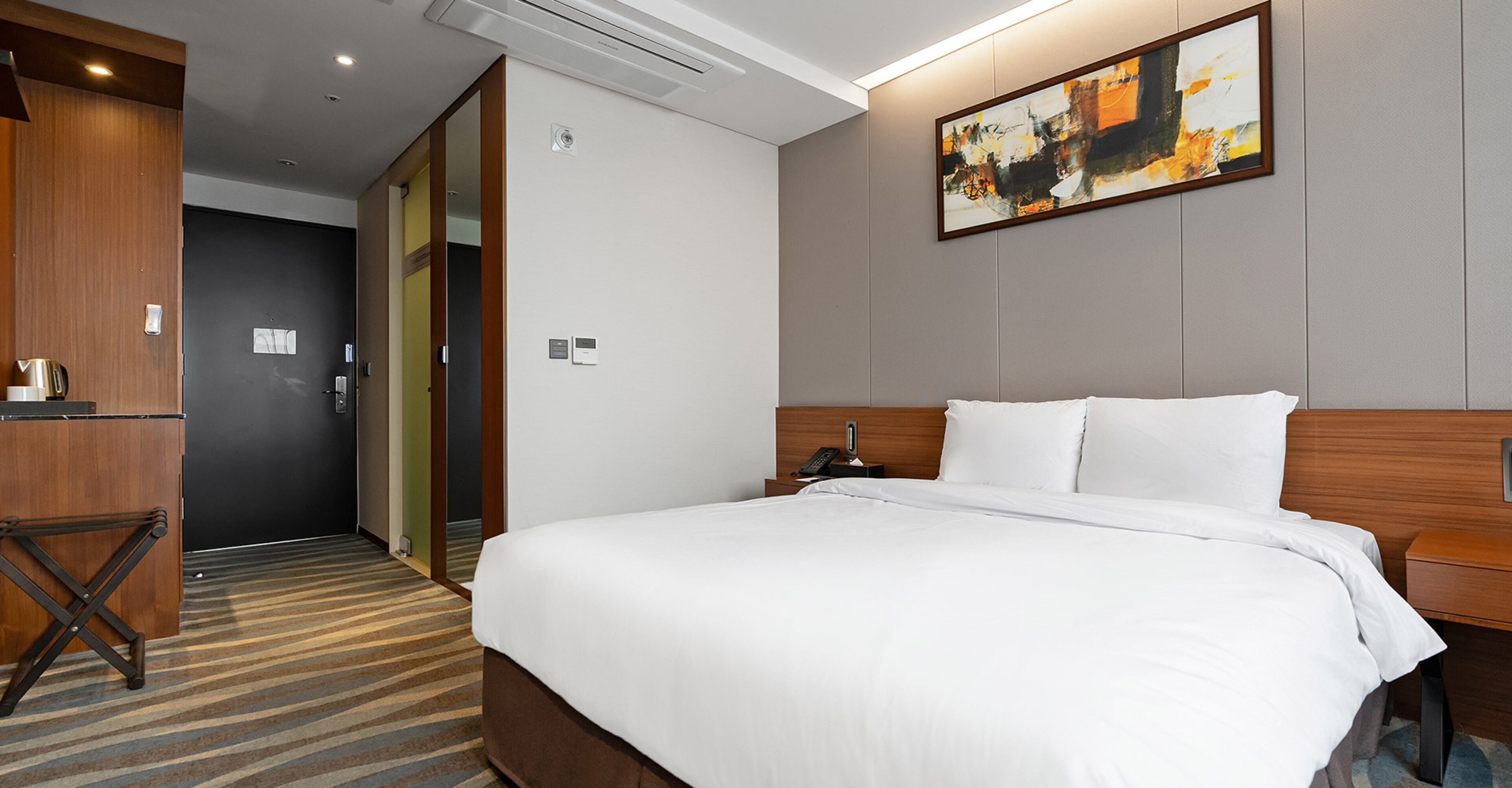 A hotel room with a large bed, a wooden headboard, a bedside table with a lamp, a painting on the wall, and a carpeted floor.