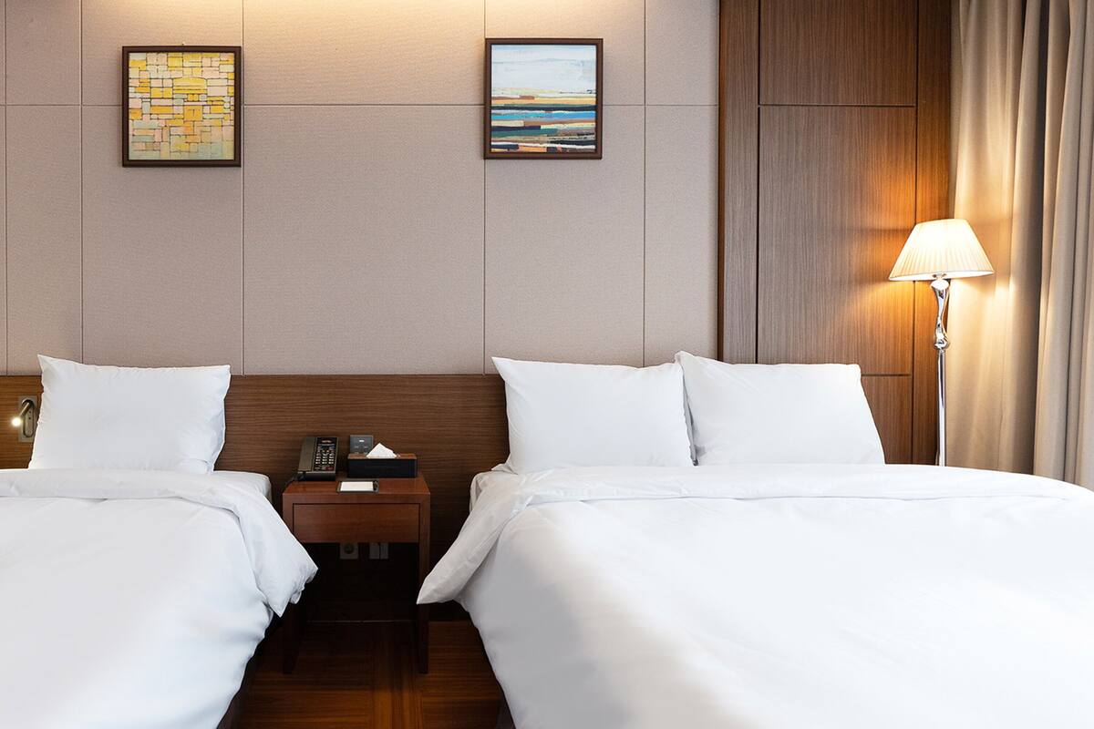 Une chambre d’hôtel avec deux lits, une tête de lit en bois, une table de chevet avec un téléphone et une boîte de mouchoirs, et deux tableaux encadrés au mur.