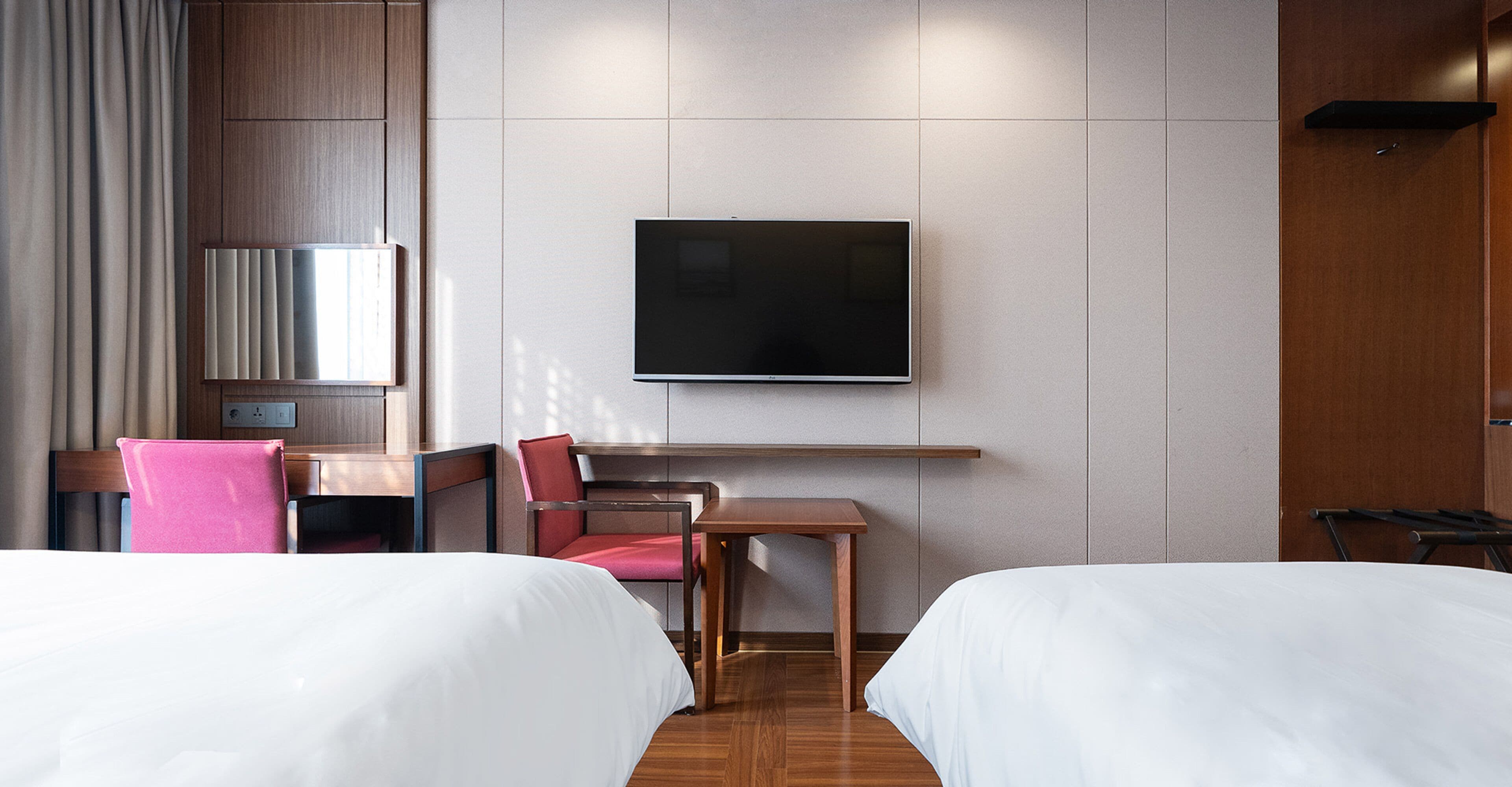 A hotel room with two beds, a desk, a chair, a television, and a wooden wardrobe.