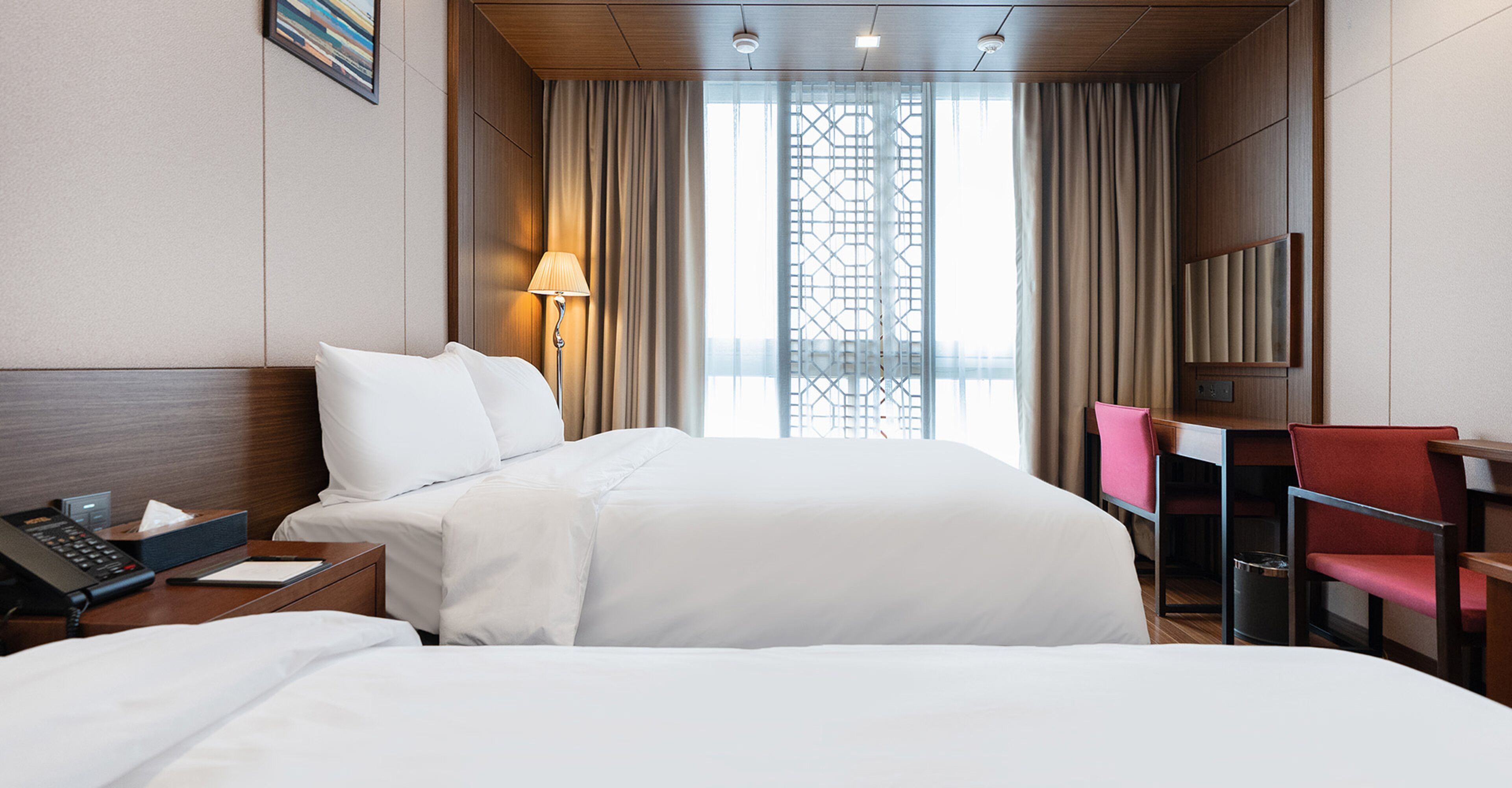 Habitación de hotel con cama, mesita de noche con lámpara, escritorio con silla y vista a un edificio a través del ventanal.