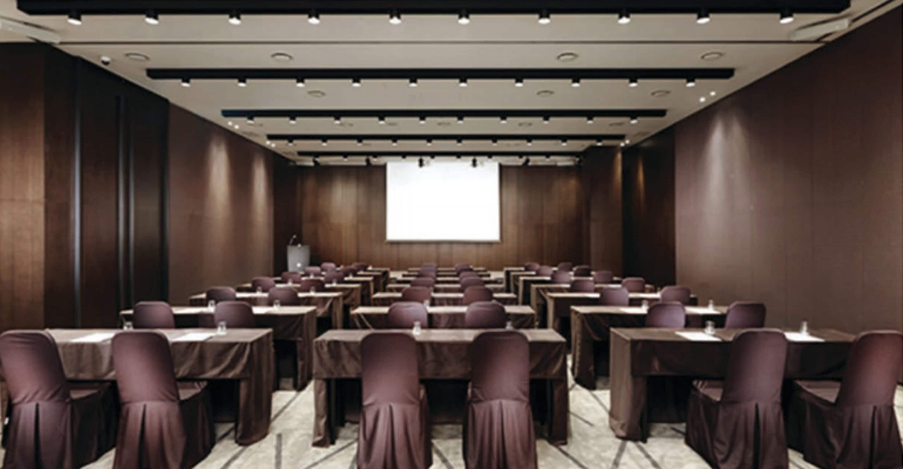 A conference room with rows of tables and chairs, a projector screen, and a podium.