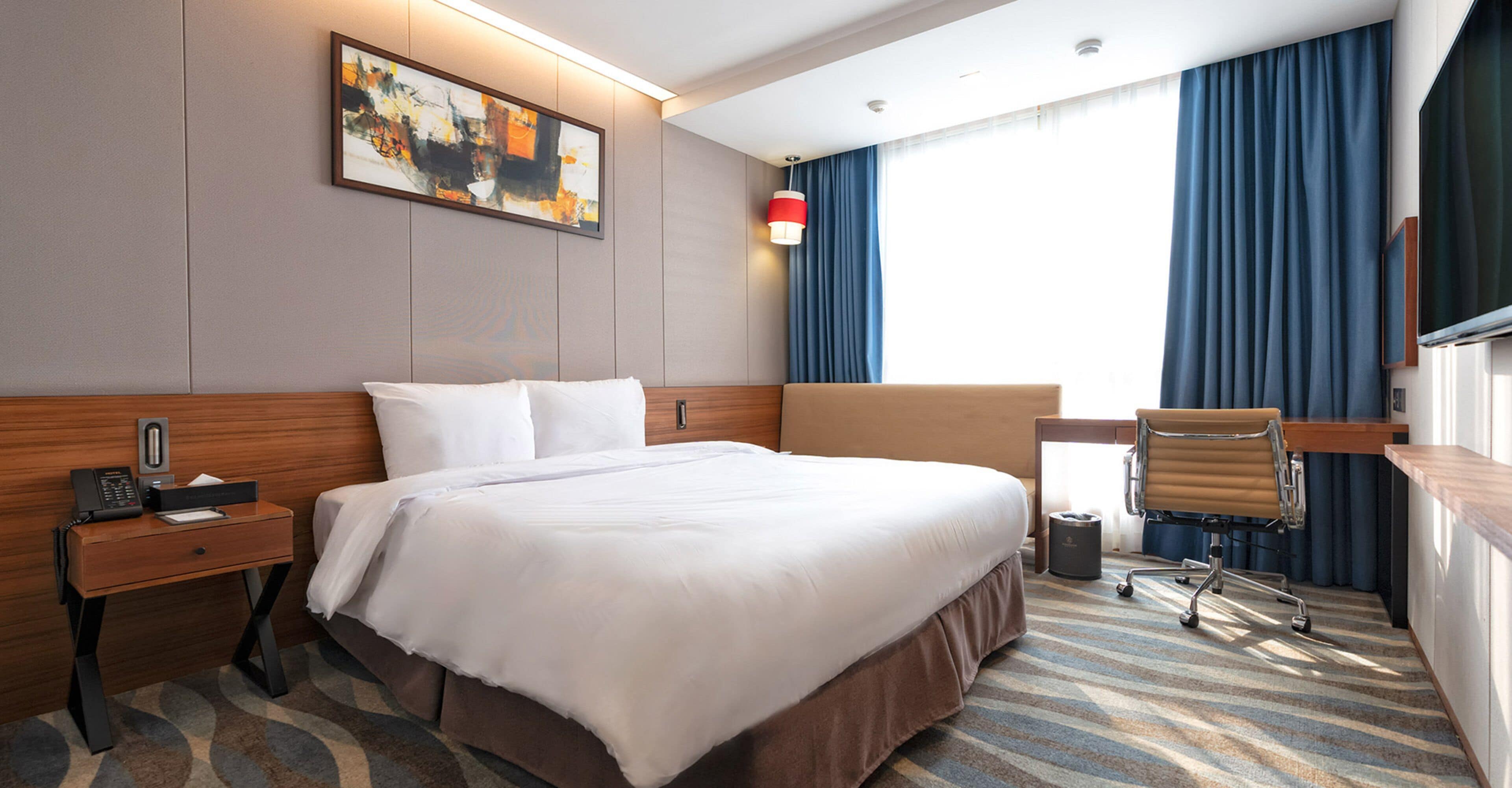 A hotel room with a large bed, a desk with a chair, a telephone, and a painting on the wall.