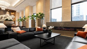 A modern hotel lobby with a black leather sofa, a coffee table, and potted plants. There is a fireplace and a large window with a city view. - Gloucesterhotel Jeonju  (Jeonju)