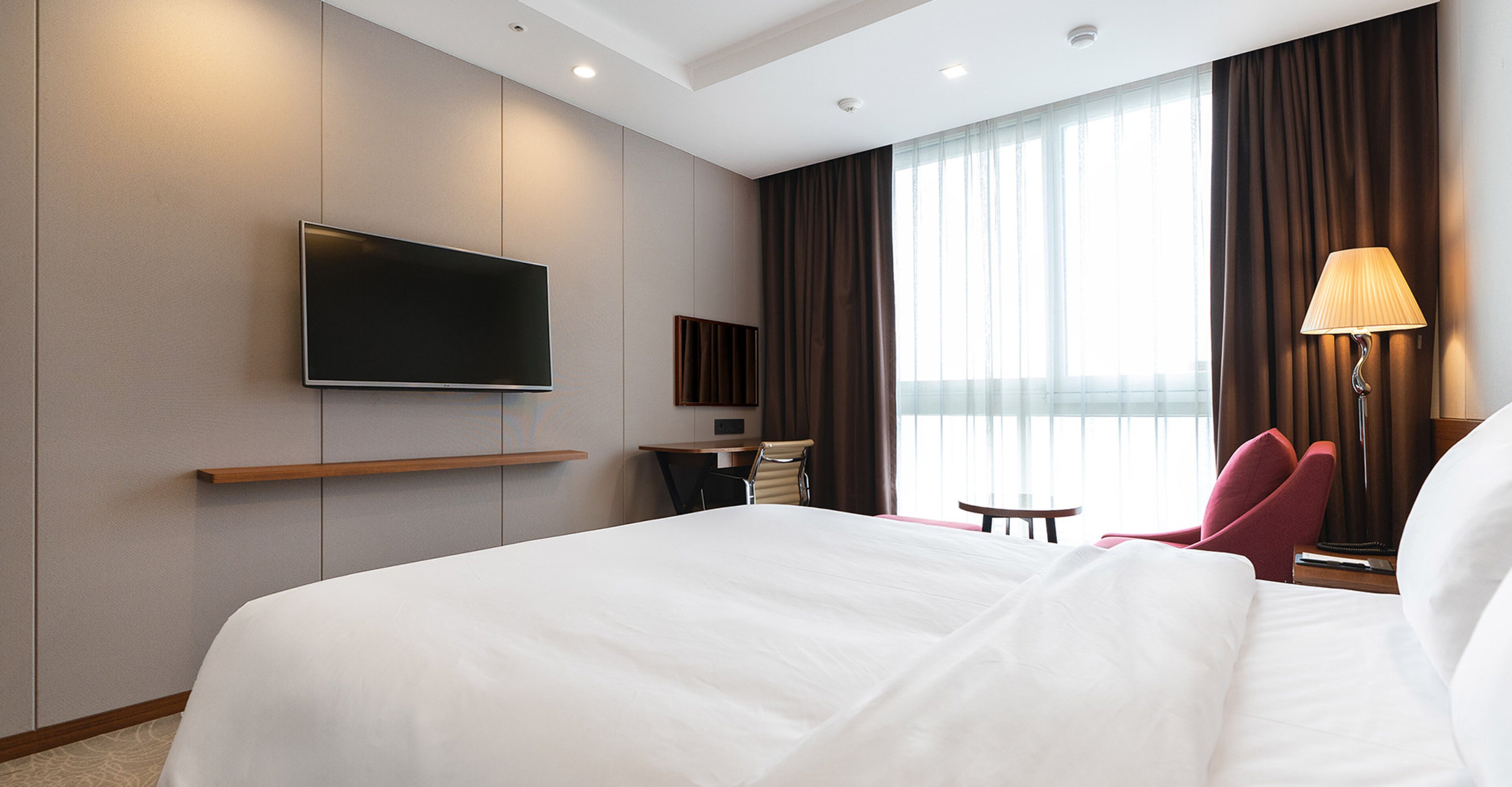 A hotel room with a large bed, a flat-screen TV mounted on the wall, a desk with a chair, a small table, and a lamp.