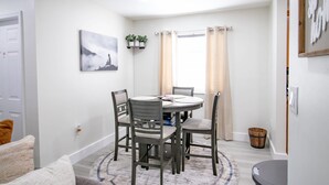 Un comedor con una mesa redonda, cuatro sillas, un cuadro, una ventana con cortinas y un pequeño estante con plantas.