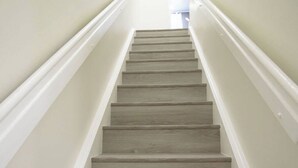 Una escalera con acabado de madera clara, paredes blancas y una lámpara en el techo.