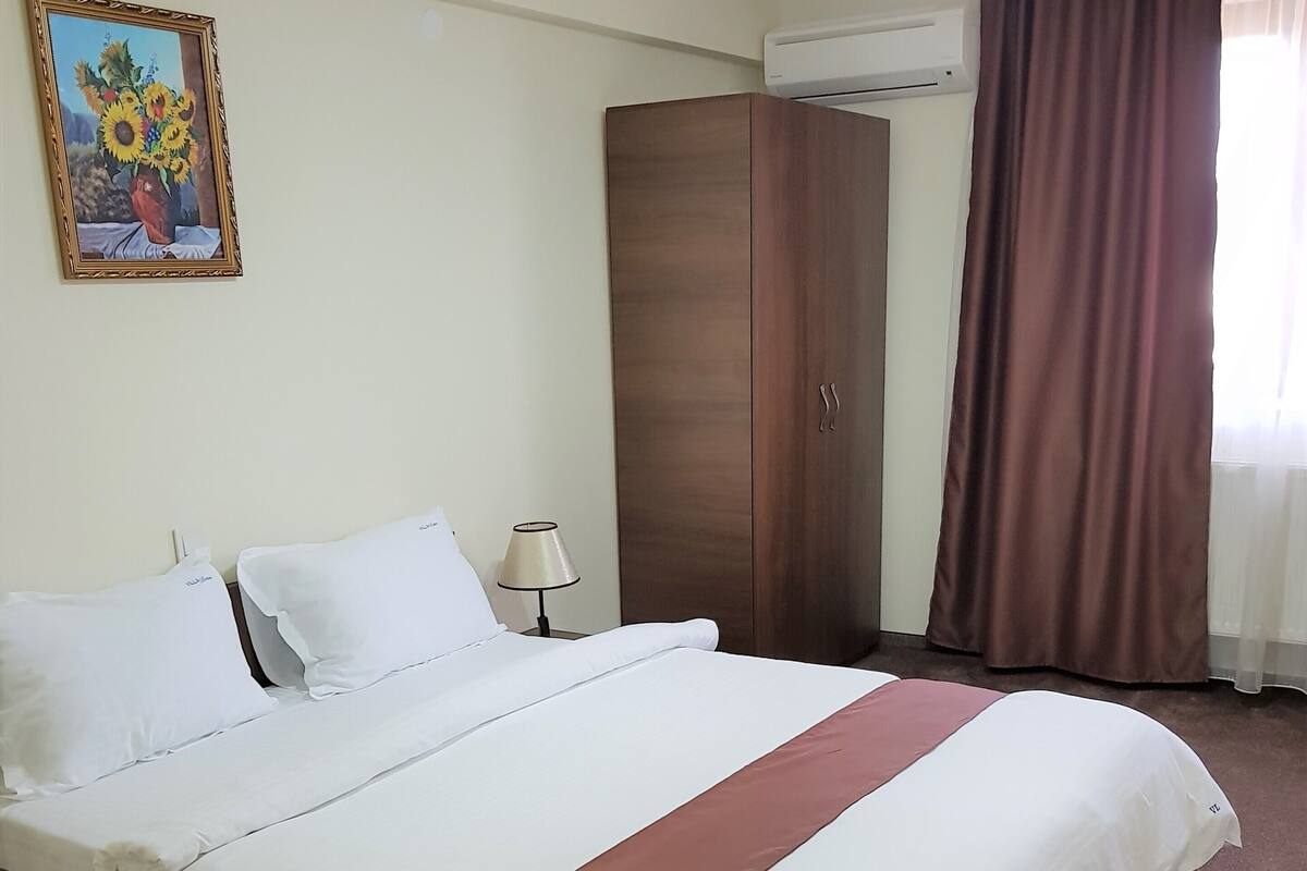 Habitación de hotel con una cama grande, un armario, un cuadro en la pared y una ventana con cortinas.